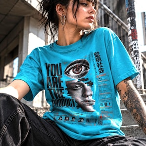 Op de afbeelding: Een felcyaan t-shirt met een grafische print met de tekst "YOU ARE THE PRODUCT", een realistisch menselijk oog, een gedeeltelijk gezicht en diverse technische datablokken met Japanse karakters en digitale interface-elementen.