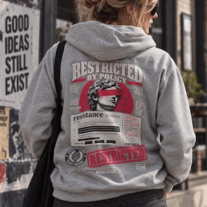 Può includere: Una felpa con cappuccio grigio melange con una grafica sul retro. Il design include la testa di una statua classica con una barra rossa sugli occhi, il testo RESTRICTED BY POLICY e una definizione da dizionario per la parola resistance.