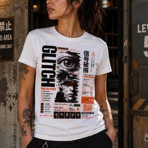 Op de afbeelding: Een wit t-shirt met korte mouwen en een grafische print met de tekst GLITCH en Reality has crashed. Het ontwerp bevat een realistisch oog, een gefragmenteerd gezicht, Japanse tekens, rode en zwarte digitale foutpictogrammen en een QR-code.