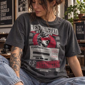 May include: A charcoal grey t-shirt featuring a graphic print of a classical statue head with a red band over the eyes. The design includes the text RESTRICTED BY POLICY, resistance, and various administrative details, including a barcode and date.