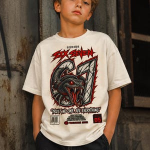 Può includere: Una t-shirt a maniche corte color bianco sporco con la grafica di un serpente nero con occhi e zanne rosse avvolto attorno al numero 67. Il testo recita SERIES SIX SEVEN, TRUST NO ONE. SEE EVERYTHING e VOL 05, con un codice a barre e testo.