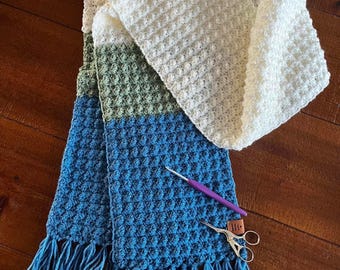 Puede incluir: Una bufanda de ganchillo hecha a mano en tonos blanco, verde y azul, con un patrón texturizado y flecos. Un ganchillo morado y unas tijeras pequeñas descansan sobre la bufanda. La bufanda se muestra sobre una superficie de madera.