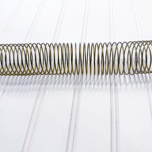 Könnte beinhalten: Eine lange, spiralförmige Metallfeder in Goldfarbe. Die Feder besteht aus einer Reihe von verbundenen Schlaufen. Die Feder liegt horizontal auf einer weißen, geriffelten Oberfläche.