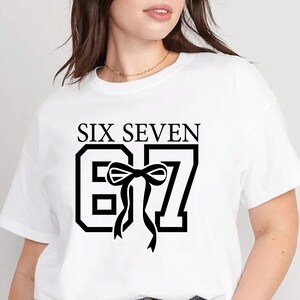K&ouml;nnte beinhalten: Wei&szlig;es T-Shirt mit dem Text "SIX SEVEN" &uuml;ber einer stilisierten "67" mit einer schwarzen Schleifengrafik. Das Design ist mittig auf der Vorderseite. Das T-Shirt hat einen Rundhalsausschnitt und kurze &Auml;rmel.