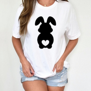 May include: White t-shirt featuring a black silhouette of a bunny with a heart-shaped detail. The bunny design is centered on the front of the shirt. The shirt has short sleeves and a crew neck.
