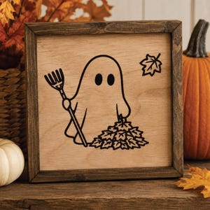 May include: Wooden framed sign with a cartoon ghost raking leaves. The ghost has two black eyes and is holding a rake. A leaf is in the upper right corner. The frame is dark brown. The background is a light wood color.