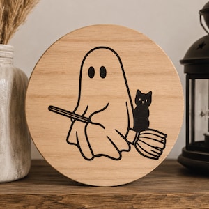 May include: A round wooden sign featuring a black illustration of a ghost riding a broom with a black cat. The ghost has two oval eyes. The sign is made of light-colored wood and is suitable for Halloween decor.