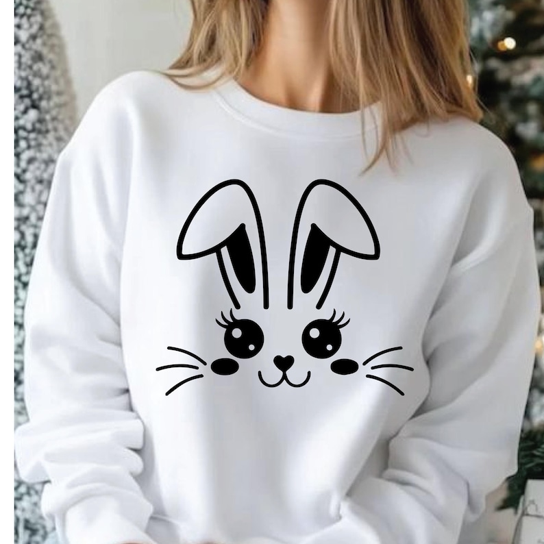 May include: White sweatshirt featuring a black bunny face design. The graphic includes large eyes with eyelashes, a heart-shaped nose, and whiskers. The bunny ears are outlined in black. A casual, comfortable top for spring or Easter.