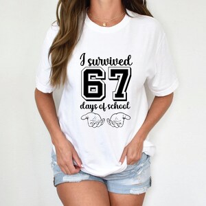 K&ouml;nnte beinhalten: Wei&szlig;es T-Shirt mit dem schwarzen Text "I survived 67 days of school". Die Zahl "67" ist fett und im College-Stil gehalten. Unter dem Text befinden sich zwei stilisierte H&auml;nde. Ein l&auml;ssiges Kleidungsst&uuml;ck f&uuml;r den Alltag.