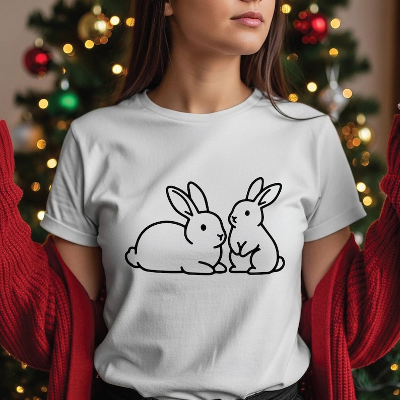 May include: White t-shirt featuring a black outline illustration of two rabbits. One rabbit is lying down, and the other is sitting upright. The shirt has short sleeves and a crew neck.