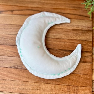 May include: A crescent moon-shaped pillow in soft white fabric with light green stitching around the edges. The pillow has a puffy, stuffed appearance and is placed on a wooden surface.
