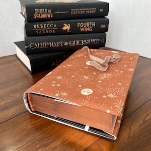 Pode incluir: Uma pilha de livros de capa dura com capas pretas e letras douradas. O livro superior tem uma capa marrom com um padrão celestial e uma fita. Os títulos incluem "Shield of Sparrows" e "Fourth Wing."
