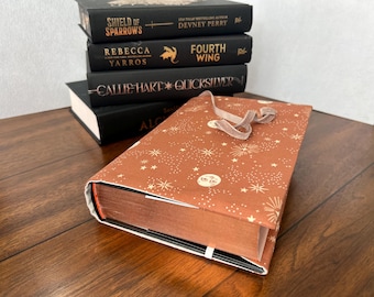 May include: A stack of hardcover books with black covers and gold lettering. The top book has a brown cover with a celestial pattern and a ribbon. Titles include "Shield of Sparrows" and "Fourth Wing."