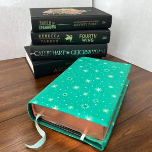 Peut inclure: Plusieurs livres à couverture rigide de couleur vert foncé. Les titres incluent "Shield of Sparrows", "Rebecca Yarros" et "Fourth Wing". Un livre turquoise avec un motif d'étoiles est au premier plan, avec un signet en ruban bleu clair.