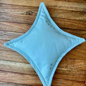 May include: A light blue, star-shaped decorative pillow with embroidered accents. The pillow has a stitched border and is placed on a wooden surface. The star shape has four points and is filled with a soft material.