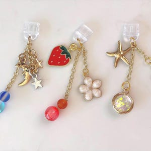 May include: Three pairs of clip-on earrings with gold chains and charms. One pair features a moon, stars, and blue beads. Another has a red strawberry and orange beads. The third has a starfish, a shell, and a round charm.