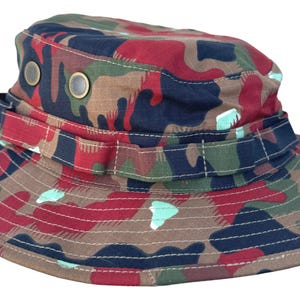 Könnte beinhalten: Camouflage-Eimerhut in einer Mischung aus Farben wie Braun, Rot, Grün und Marineblau. Der Hut hat eine breite Krempe und einen Riemen um den Kopf. Zwei Metallösen sind an der Seite sichtbar.