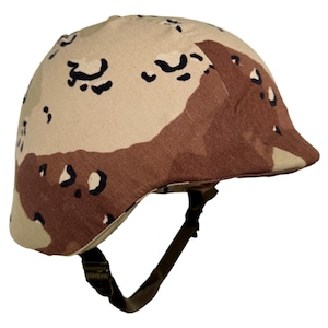 Könnte beinhalten: Ein tarnfarbener Helm in Beige- und Brauntönen mit Kinnriemen. Der Helm hat eine abgerundete Form und besteht aus einem strapazierfähigen Material. Das Tarnmuster besteht aus unregelmäßigen Formen in verschiedenen Braun- und Beigetönen.