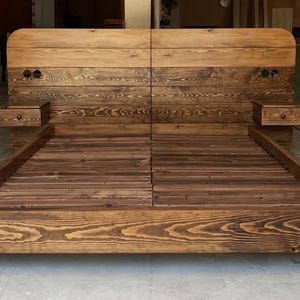 Peut inclure: Un lit en bois fait à la main avec une tête de lit et deux tables de chevet intégrées. Le lit est en bois teinté avec des veines apparentes. Les coins sont renforcés par des équerres métalliques noires. La tête de lit a un dessus incurvé.