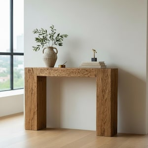 Puede incluir: Una consola rectangular de madera con acabado natural. La mesa tiene gruesas patas en forma de bloque y una tapa a juego. Un jarrón con vegetación, libros y objetos decorativos se encuentran sobre la mesa. La mesa está en un salón moderno.