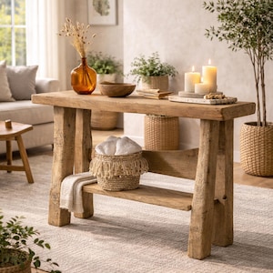 Könnte beinhalten: Ein rustikaler Holztisch mit einer unteren Ablage, auf der sich ein geflochtener Korb mit Fransen und ein gefaltetes Handtuch befinden. Der Tisch ist mit Kerzen, einer Holzschale und einer Vase mit getrockneten Blumen geschmückt. Der Raum hat eine neutrale Farbpalette.