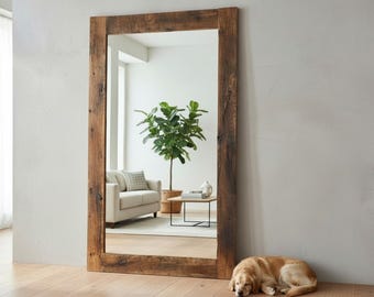 May include: A large rectangular mirror with a rustic, weathered wooden frame. The mirror reflects a living room scene with a sofa, plant, and table. A golden retriever dog is lying on the wooden floor in front of the mirror.