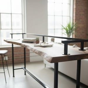 May include: A live edge wooden desk with black metal legs and a black metal railing. The desk has books, glasses, and a small plant on top. A gray stool sits to the left. The room has large windows and a brick wall.