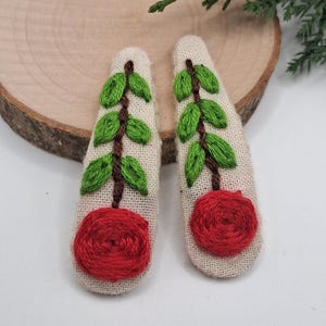 May include: Two fabric hair clips with embroidered designs. Each clip features a red circular shape at the bottom, a brown stem, and green leaves. The clips are on a light-colored fabric.