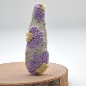 May include: Embroidered fabric bottle stopper with three purple and beige flower designs. The stopper is made of a natural-coloured burlap-like material and is displayed on a wooden slice. The flowers have a textured, handmade appearance.