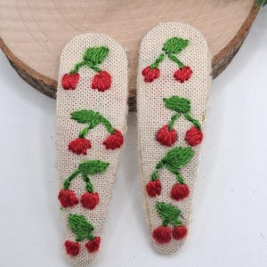 May include: Two embroidered hair clips with a cherry design. Each clip features a beige fabric base with red cherries and green leaves. The clips are approximately 3 inches long.