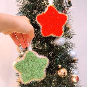 May include: Two star-shaped Christmas decorations, one red and one green, hang on a small Christmas tree. The red star has a glittery texture, while the green star has a fuzzy texture. Both stars have a white and pink trim. The tree is decorated with silver and gold baubles.