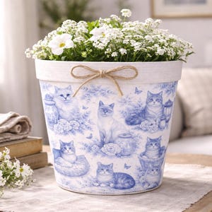 May include: A white tapered flower pot featuring a blue toile-style pattern of various cats, flowers, and jars. The rim is textured white with a tied jute twine bow at the center. The pot is filled with small white flowers and green foliage.