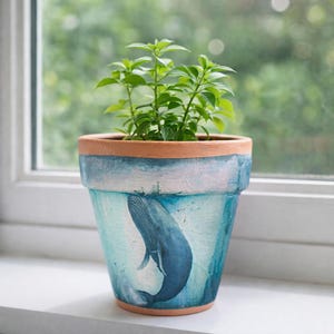 May include: A terracotta plant pot with a blue whale design. The pot has a light blue and white painted background. The pot is filled with a green leafy plant. The pot is sitting on a white windowsill.