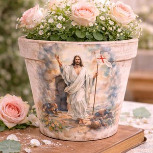 May include: A beige ceramic flower pot with a painted image of Jesus Christ holding a flag, filled with pink roses and white baby's breath. The pot sits on a wooden book.