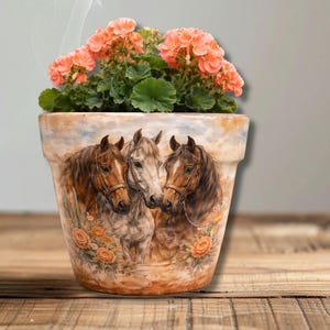 May include: A decorative terracotta flower pot featuring a painted image of three horses. The pot is filled with green foliage and vibrant orange flowers. The pot is sitting on a wooden surface.