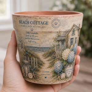 May include: A terracotta plant pot featuring a printed beach cottage scene with blue and white hydrangeas, a lantern, and seashells. The text reads BEACH COTTAGE, SIMPLE JOYS BY THE SEA, Collect seashells, soak up the sun, and let the worries drift away.