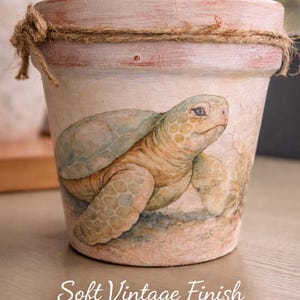 May include: A decorative terracotta flower pot with a painted sea turtle design. The pot has a soft vintage finish with muted, matte colors. A twine rope is wrapped around the rim. The text on the pot reads "Soft Vintage Finish Muted, Matte Colors."