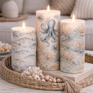 May include: Three lit pillar candles with ocean-themed designs. One candle features fish, another an octopus, and the third coral. The candles are on a woven tray with decorative coral and beads. The background includes a neutral-toned sofa and pillows.