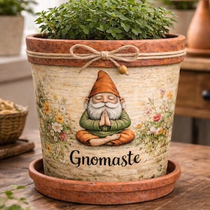May include: A terracotta-colored plant pot with a matching saucer, featuring a cream-colored body with a printed illustration of a gnome meditating in a lotus position, surrounded by wildflowers, with the text Gnomaste and a jute twine bow.