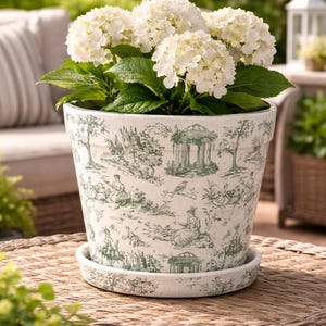 May include: A white ceramic planter with a green toile design, filled with white hydrangea flowers. The planter sits on a matching saucer. The background includes a wicker table and outdoor seating.