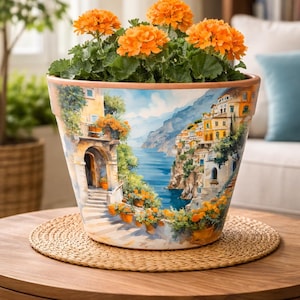 May include: A decorative ceramic flower pot featuring a painted coastal scene with a blue sea and colorful buildings. The pot is filled with vibrant orange flowers and green foliage. The pot sits on a round wooden table.