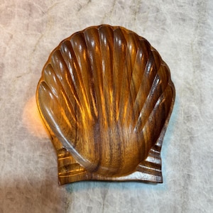Puede incluir: Un cuenco de madera con forma de concha con un acabado de vetas de madera natural. El cuenco tiene un borde festoneado y un interior poco profundo. El cuenco mide aproximadamente 15 cm de ancho y 13 cm de largo.