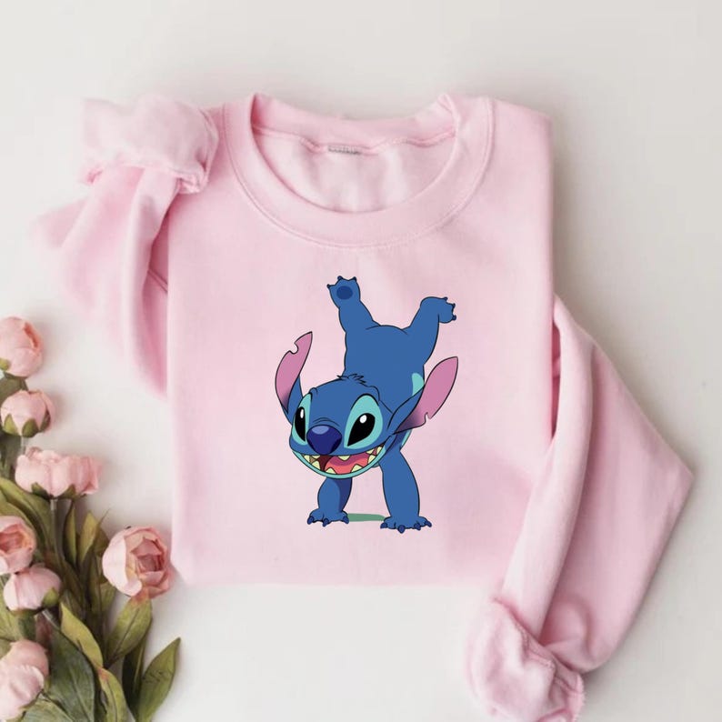 May include: A light pink sweatshirt featuring a blue cartoon character, Stitch, in a playful pose. The character has large ears, a wide smile, and is set against a plain background. The sweatshirt has long sleeves.