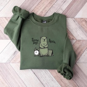 May include: Olive green sweatshirt with a graphic of a groundhog, alarm clock, and calendar. The text on the sweatshirt reads "Same Day. Again.". The groundhog is drawn in a cartoon style.