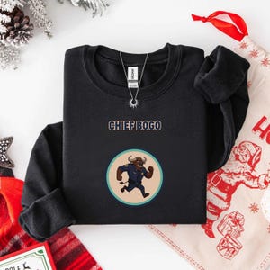 May include: Black crewneck sweatshirt with the text "CHIEF BOGO" above a circular graphic of a cartoon bull in a police uniform. The bull is running. A silver necklace is on the sweatshirt.