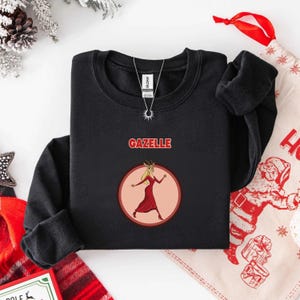 May include: Black sweatshirt with the word "GAZELLE" in red block letters above a circular graphic. The graphic features a cartoon gazelle in a red dress. A silver necklace with a sun pendant is visible.
