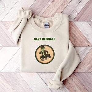 May include: A cream-coloured sweatshirt featuring the text "GARY DE'SNAKE" above a circular graphic. The graphic depicts a cartoon snake in a hat and coat. The sweatshirt is folded on a wooden surface.