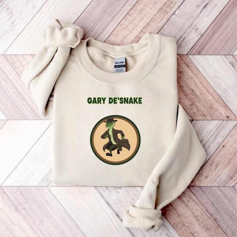 May include: A cream-coloured sweatshirt featuring the text "GARY DE'SNAKE" above a circular graphic. The graphic depicts a cartoon snake in a hat and coat. The sweatshirt is folded on a wooden surface.