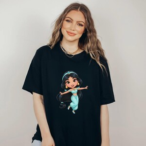 Puede incluir: Camiseta negra con una ilustraci&oacute;n de dibujos animados de la Princesa Jasmine con un atuendo turquesa, diadema a juego y pendientes dorados. La princesa tiene el pelo largo y negro y est&aacute; sonriendo. La camiseta la lleva una persona.