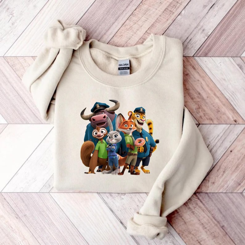 Puede incluir: Una sudadera de color crema con un gr&aacute;fico colorido de personajes de la pel&iacute;cula animada Zootopia. Los personajes incluyen un b&uacute;falo, una ardilla, un conejo, un zorro y un tigre, todos con uniformes de polic&iacute;a. La sudadera est&aacute; doblada sobre una superficie de madera.